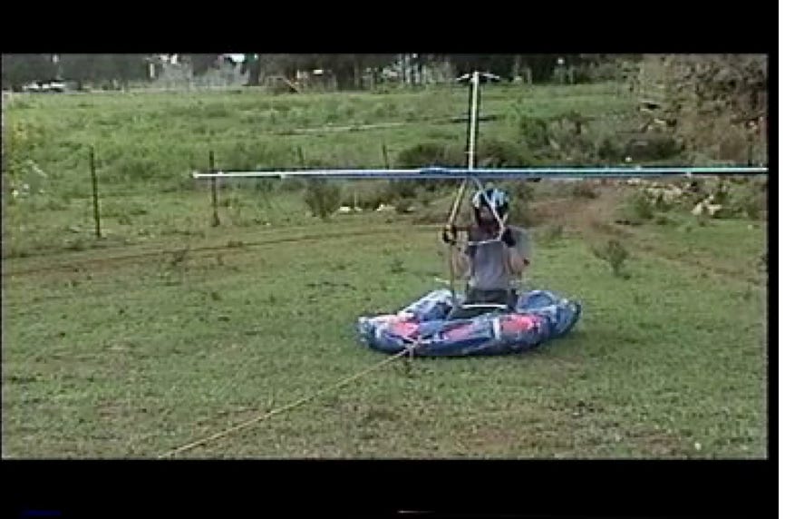 A grown man sitting in a children's swimming pool that has been wrapped in a blue tarp, while wearing a motorcycle helmet and holding on to a homemade hang glider.