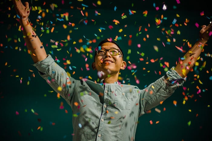 Androgynous Asian person smiling and standing with arms outstretched as colourful confetti falls around them