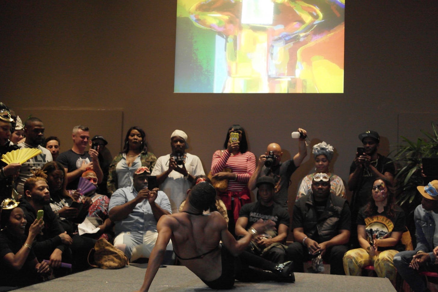 Photo taken from behind a ballroom performer seated at the end of a stage in a dance pose. An audience of primarily Black people with some dressed in ballroom attire watches attentively.