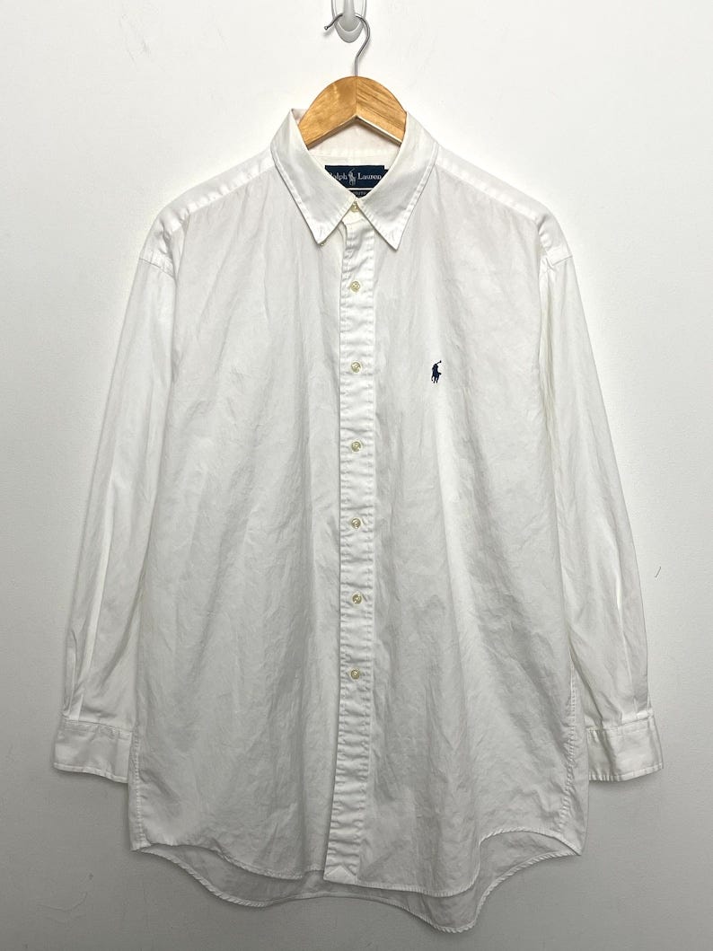 May include: A long-sleeve, white button-down shirt with a small embroidered logo on the chest. The shirt is made of a lightweight cotton material and has a classic, relaxed fit.  The shirt is shown hanging on a wooden hanger against a white background. The Polo Ralph Lauren label is visible on the inside of the collar.