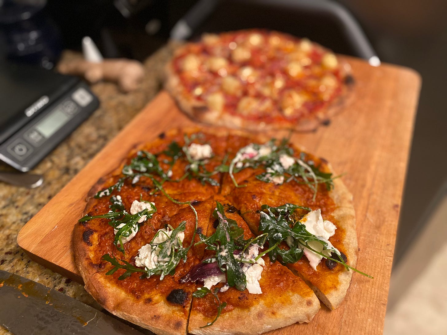 Two pizzas on the cutting board. In the foreground is the one with vodka sauce, burrata, and arugula, as described above. Blurred in the background is a capocollo pineapple one.