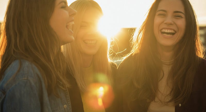 Prompt: Three women stand together laughing, with one woman slightly out of focus in the foreground. The sun is setting behind the women, creating a lens flare and a warm glow that highlights their hair and creates a bokeh effect in the background. The photography style is candid and captures a genuine moment of connection and happiness between friends. The warm light of the golden hour lends a nostalgic and intimate feel to the image.
