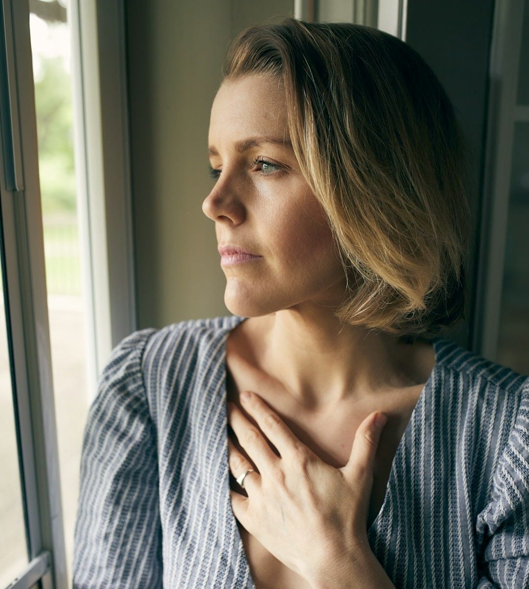 a woman looking out a window with her hand on her chest