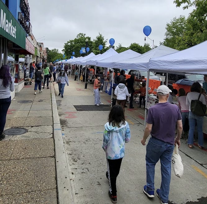 Picture shows the old service lane on the east side of Connecticut. The view is south next to Yes Organic Market, whose awning can be seen on the left. On the right, are 10-15 white tents in a line with a blue balloon on top of each. Lots of people are walking and shopping at the outdoor market.