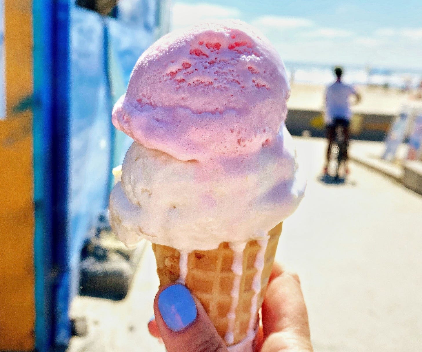 close up of a waffle cone with scoops of pink and white ice cream. cone is held by a hand with bright blue nail polish. this against a seaside background with bright blue beach shack to the left