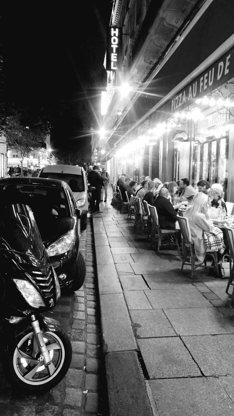 A black and white photo with a line of parked cars on the left side, and a scene of people dining outdoors at a cafe on the right side.