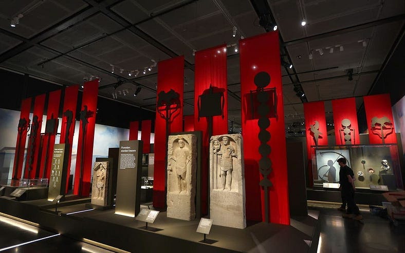 The British Museum Legion exhibition. Red strips of cloth hang like standard banners from the ceiling to the floor, with black cut out standards infront of them, almost sillhouetted. On black raised platforms are different exhibits, including stone funerary monuments to different Roman soldiers.