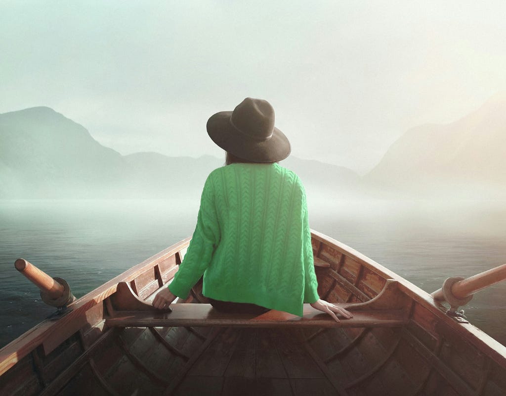 Woman in a green cardigan and dark gray felt wide brim hat sitting in a rowboat with the handles of the oars on either side pointed up. The paddle portion of oars are out of sight in the water. Her hands are resting on the boat seat on either side of her. Fog and mist hover over the dark blue gray water in front of the boat, with green mountains lining the opposite shore.