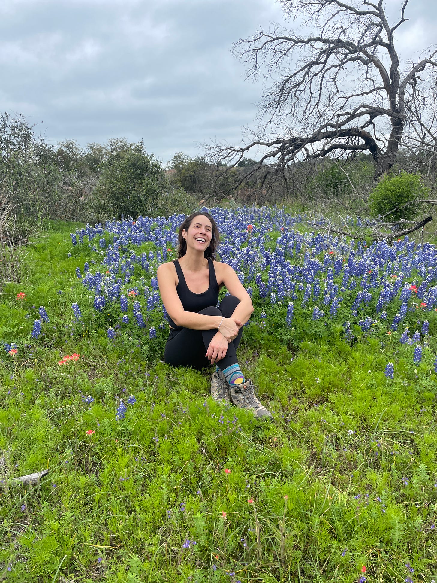 A white woman with brown hair in black athletic clothing and hiking boots sits in front of a patch of blooming bluebonnets, smiling.