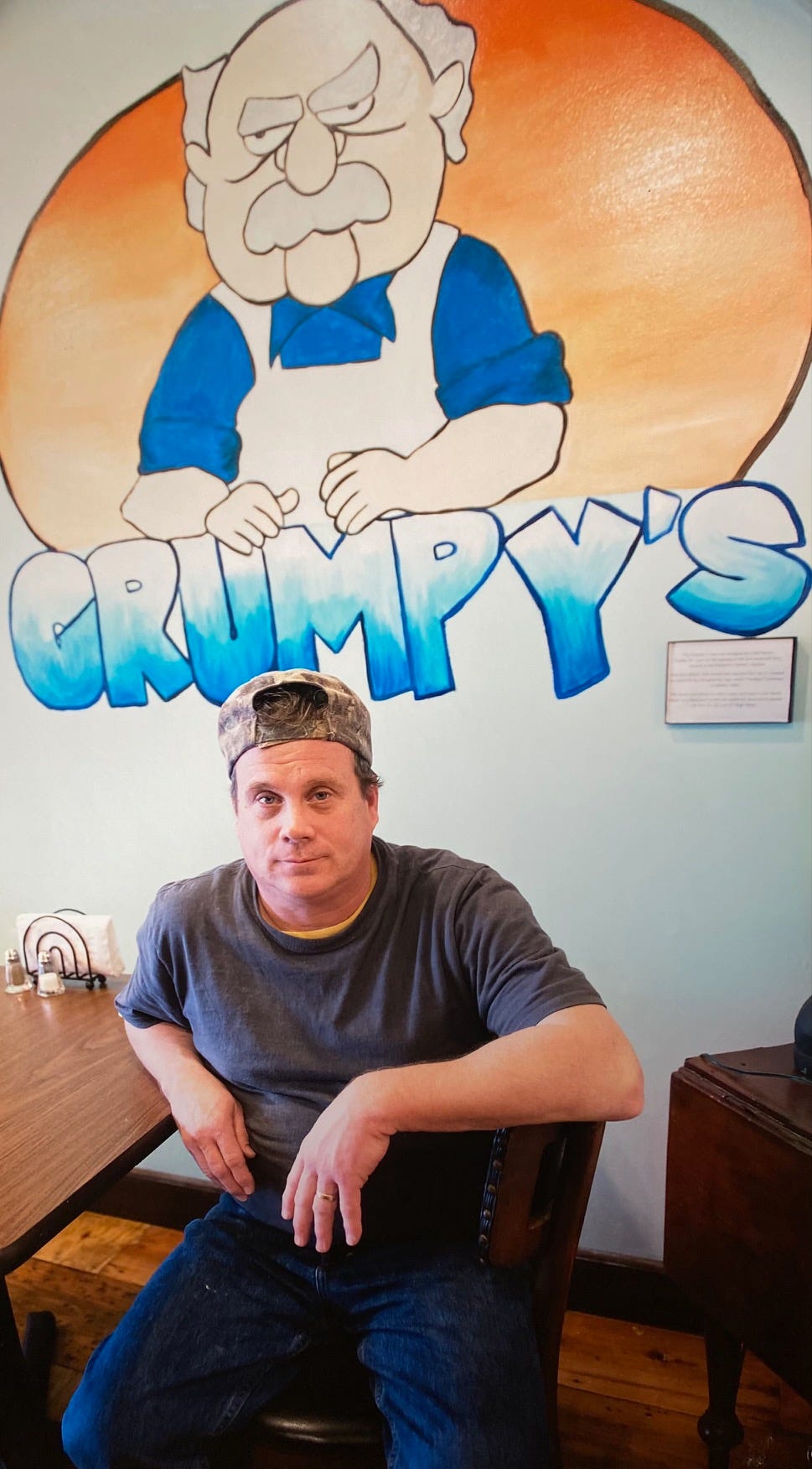 White man seated in chair, looking at camera, wearing dark t-shirt, blue jeans, and backwards camo hat with large caricature of grumpy old man painted on wall behind him and GRUMPY'S spelled out in bright blue.