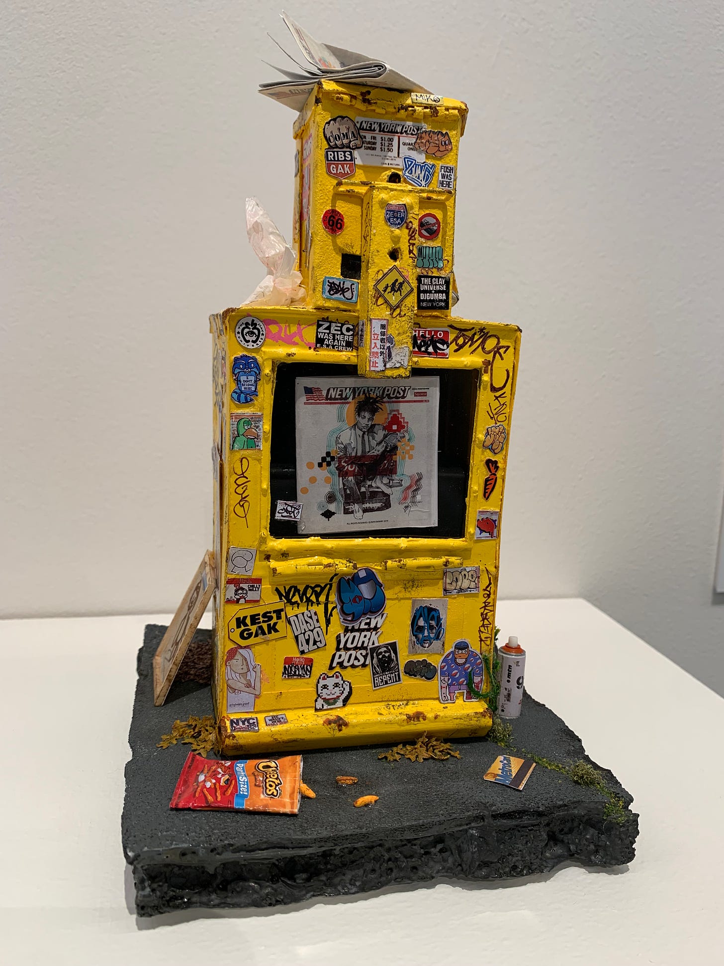 A yellow news box found on an NYC street, in miniature. It's covered with stickers and graffiti, and litter is strewn on and around it