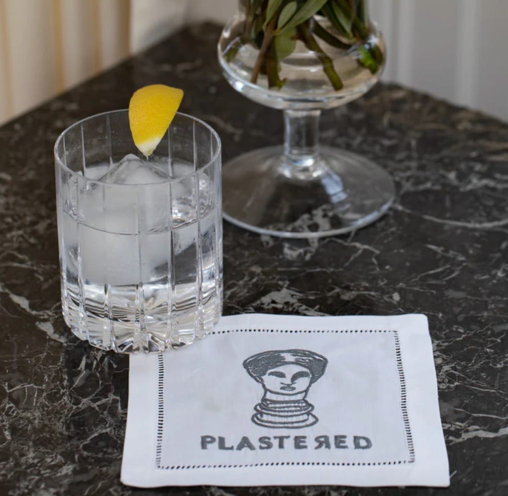 A black marble table with a clear liquid cocktail in a glass and a white square napkin with a drawing of a bust that says “PLASTERED”