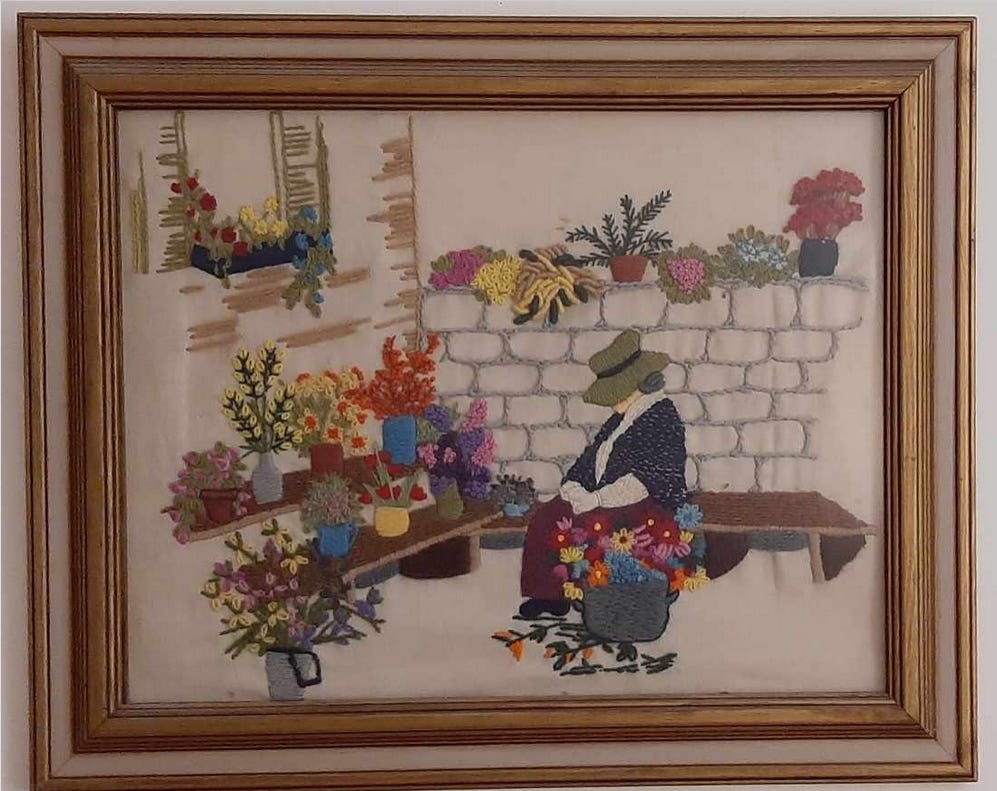 Framed needlework picture of a woman sitting outdoors making a flower arrangement with wooden benches with flower arrangements and plants and more plants on a stone wall. Framed needlework picture of a woman sitting outdoors making a flower arrangement with wooden benches with flower arrangements and plants and more plants on a stone wall.