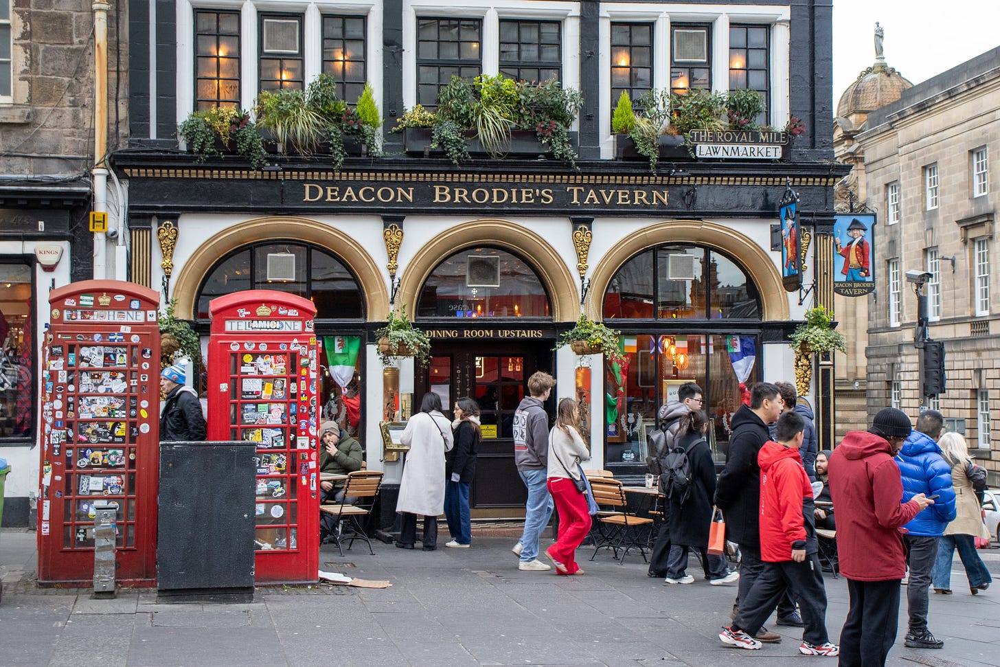 A busy street in front of the Deacon Brodies Tavern pub, on the corner of the Royal Mile. In the foreground are two red phone boxes, covered in hundreds of stickers