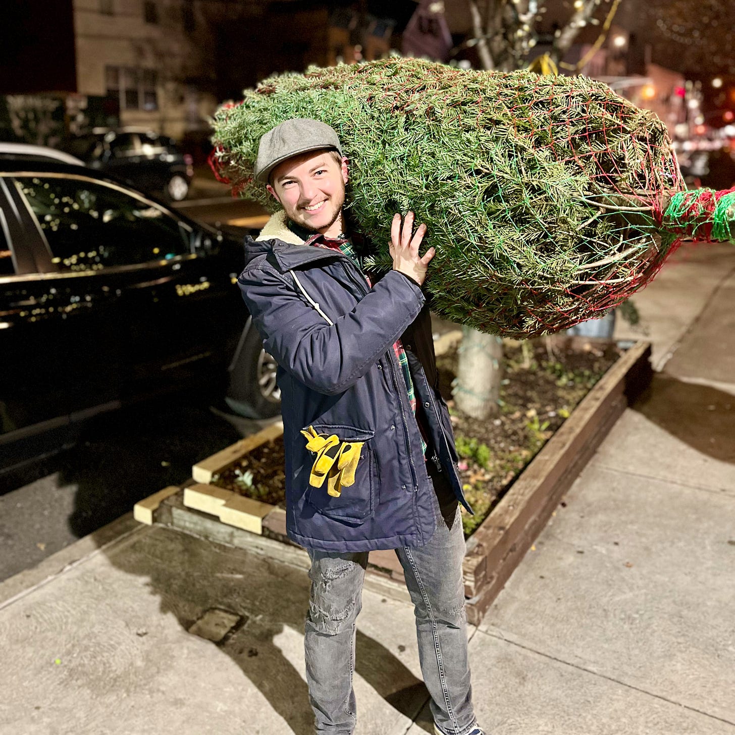Jackson Bird holding a netted Christmas tree over his shoulder on a New York City street in the evening. He's smiling broadly at the camera and dressed in a winter coat and flat cap with work gloves sticking out of his pocket.