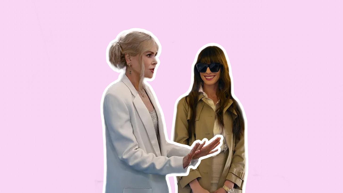 Nicole Kidman and Anne Hathaway stand against a solid pastel pink backdrop with a thick white border outlining their silhouettes. On the left, Kidman wears a white suit and with her platinum blonde hair in a bun. To her right is Hathaway, wearing large black sunglasses and a tan trenchcoat, with her brown hair down with heavy bangs.