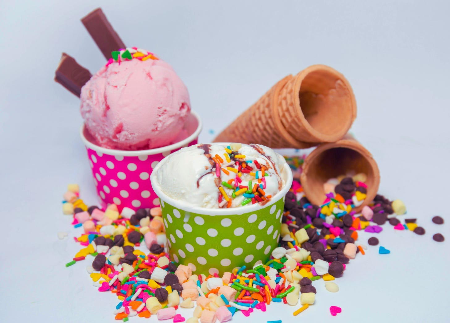 Two cups of ice cream with sprinkles and chocolate pieces, alongside empty waffle cones on a white background.