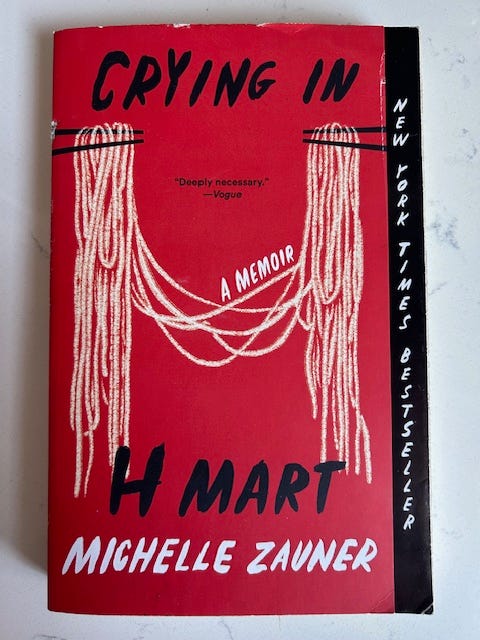 A red book cover where noodles are dangled from chopsticks into the shape of the letter H. It reads, Crying in H Mart A Memoir by Michelle Zauner, New York Times Bestseller.