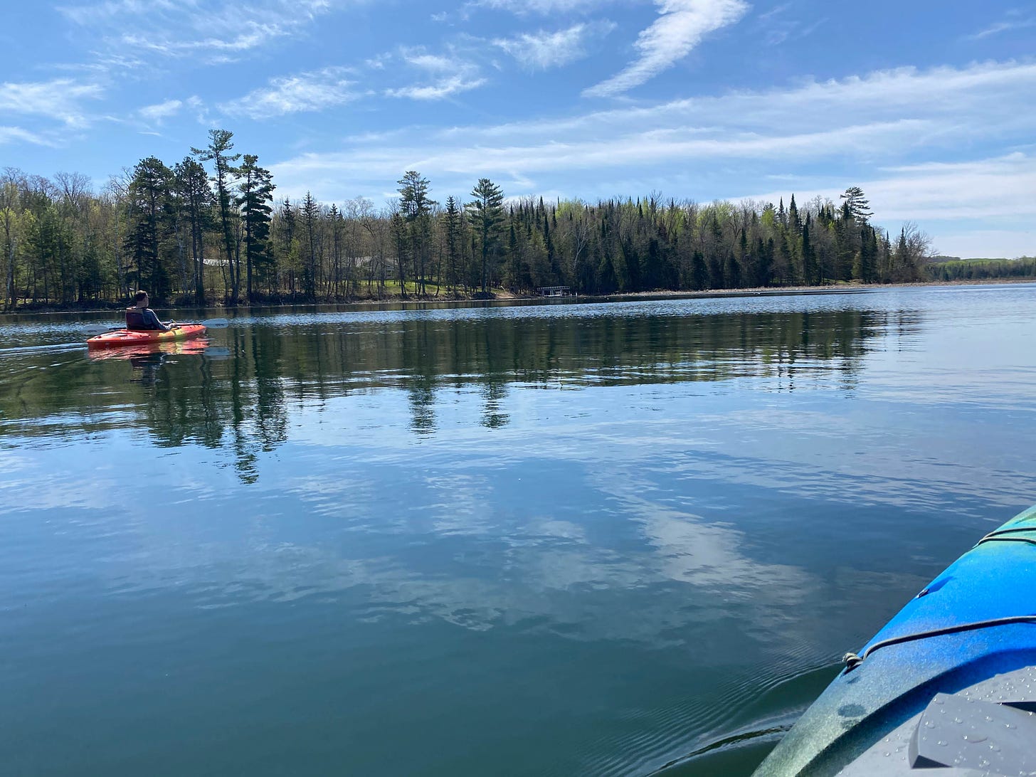 View from a kayak of another kayak on a still blue lake, reflecting white clouds and blue sky above.