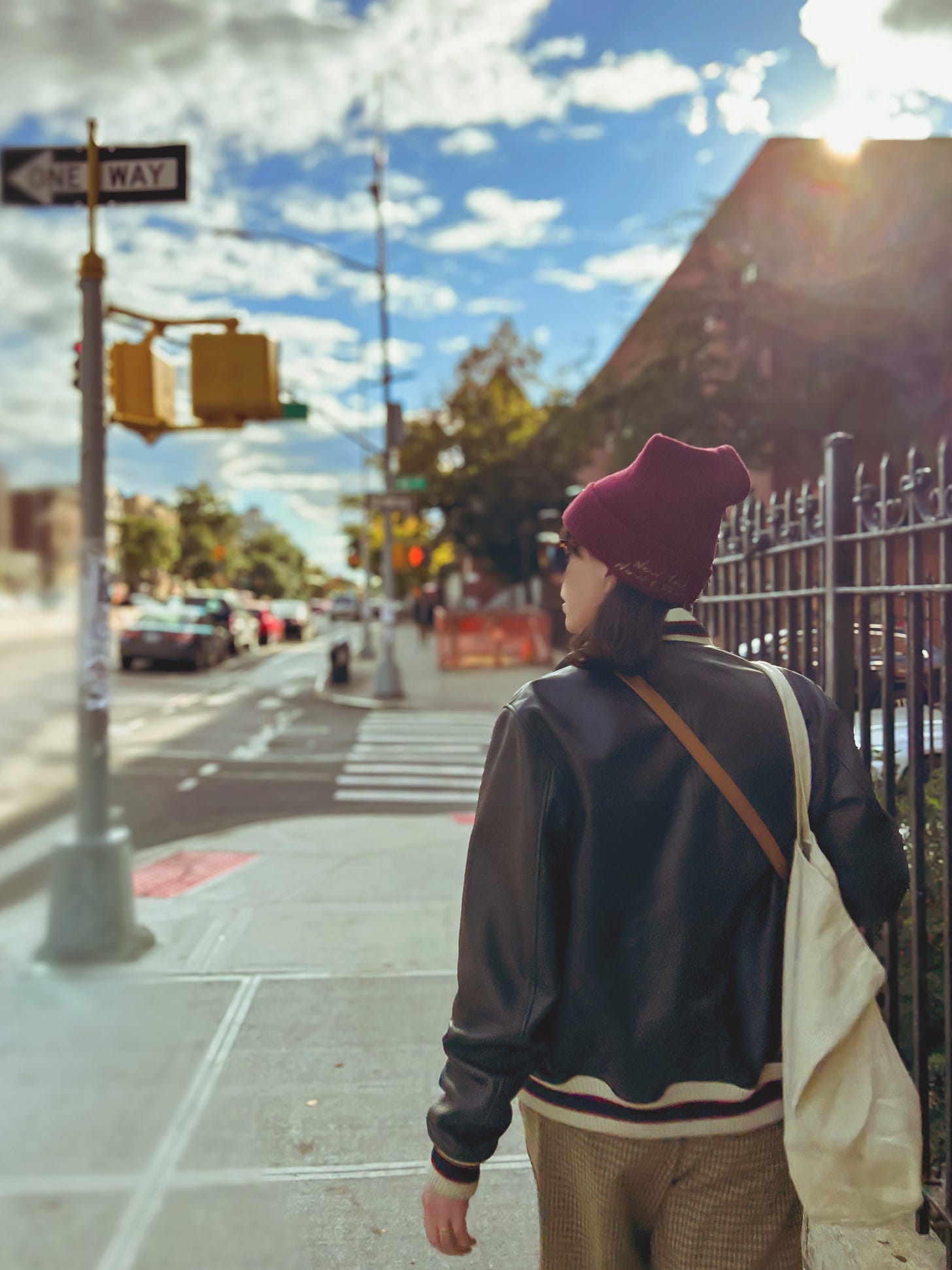 Jen, a white queer with shoulder length dark hair, walks down a Brooklyn street as the sun sets over a building. She's wearing sunglasses, a maroon beanie, a black jacket and grey pants. 