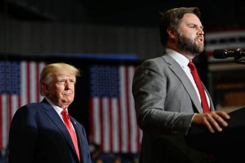 Two suited men - one elderly, the other middle-aged - on stage in front of US flags.