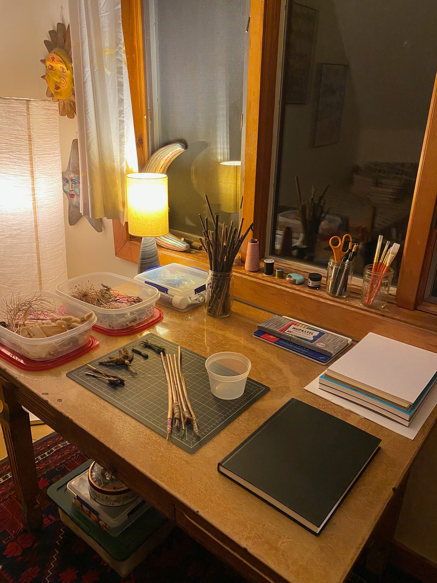 A table under a dark window, lit by a small lamp. The table and windowsill are full of stacks of paper, brushes-in-progress, containers of plant materials, colored pencils, and a my sketchbook. 