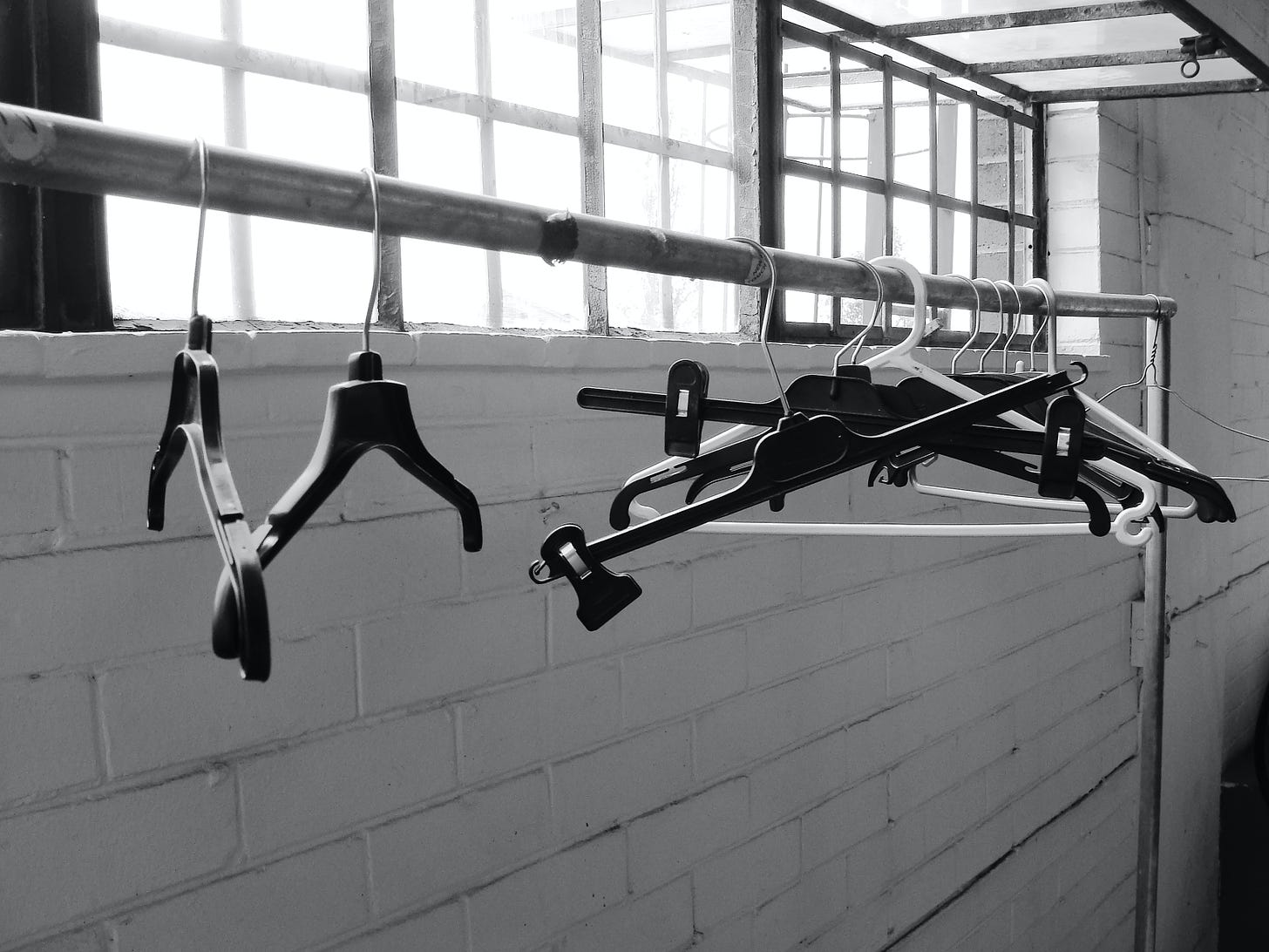 Empty coat hangers hang on a metal bar by an open window and against a brick wall