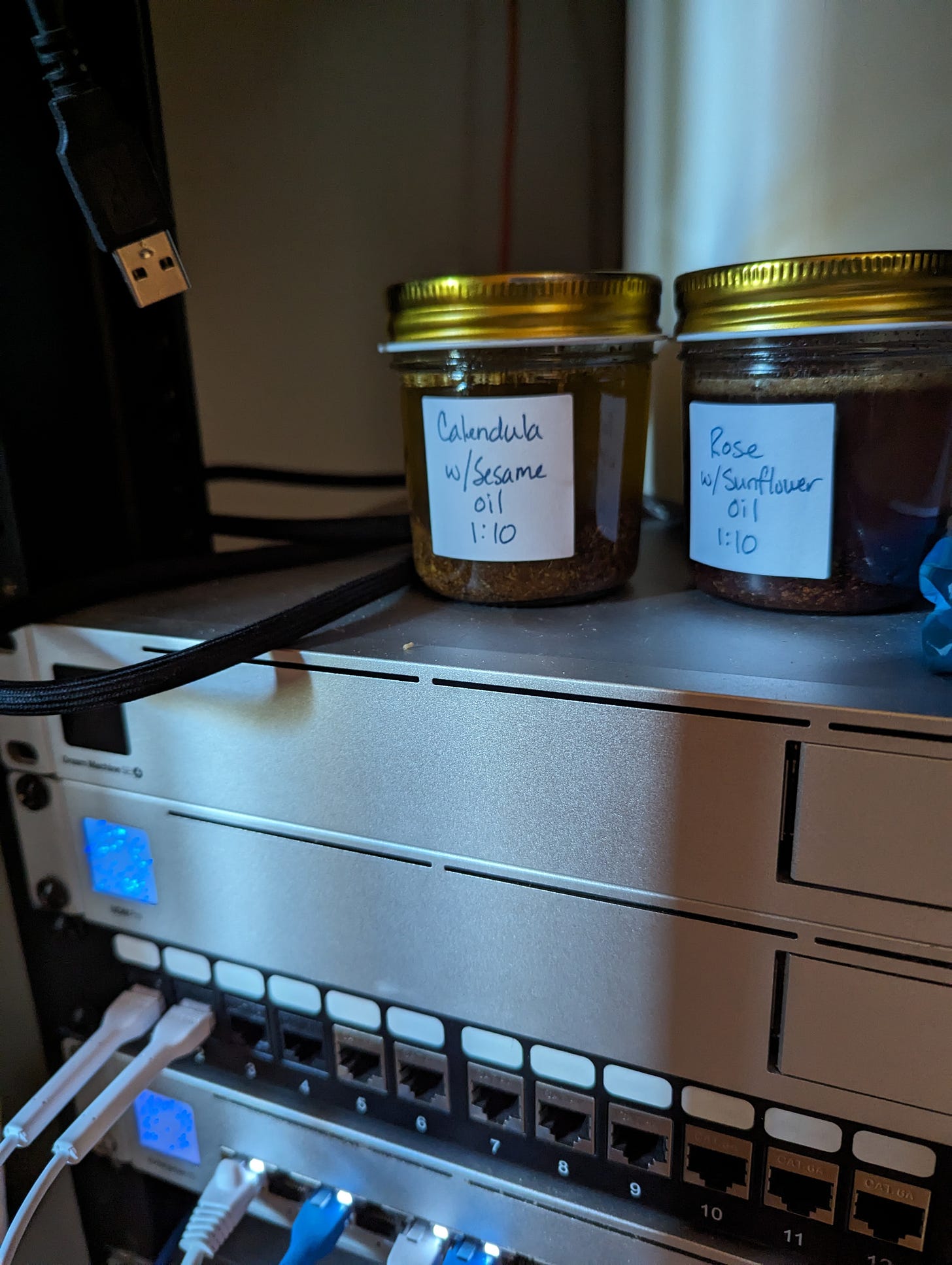 2 jars labeled "calendula withe sesame oil" and "rose with sunflower oil" rest on top of a server components. ethernet cables can be seen in some of the ports and a USB plug hangs down.