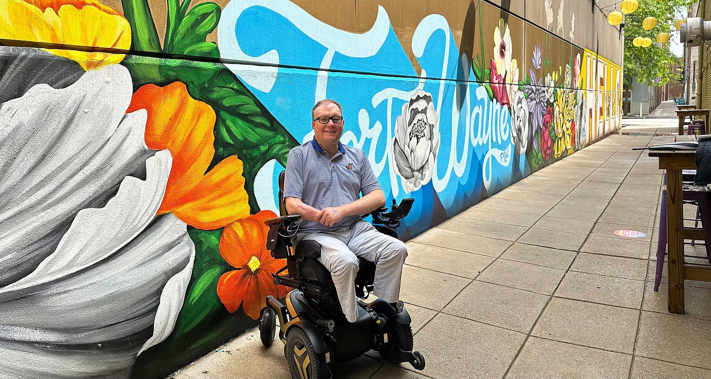 John seated in his wheelchair against the wall of an alley that is painted with a floral mural and text that reads Fort Wayne.