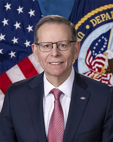 The image shows a man wearing glasses, a dark suit, a white shirt, and a red patterned tie. He is standing in front of two flags: the United States flag and a flag with the seal of the Department of Veterans Affairs. The background and setting suggest an official or government portrait.