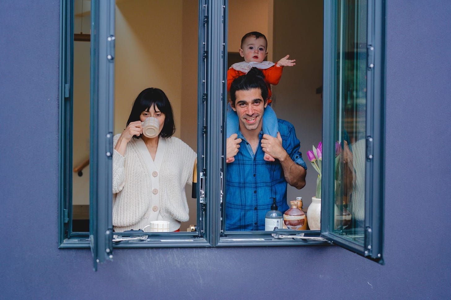 two adults looking out of a casement window. one is drinking coffee. the other is smiling and has a child on their shoulders. the child is waving