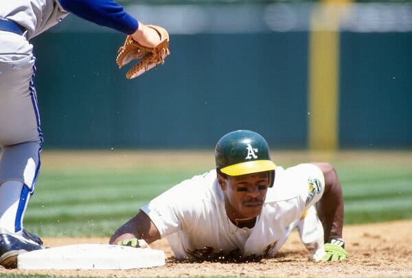 Rickey Henderson winces as he slides headfirst during a baseball game. He's wearing a green and yellow Oakland A's helmet.