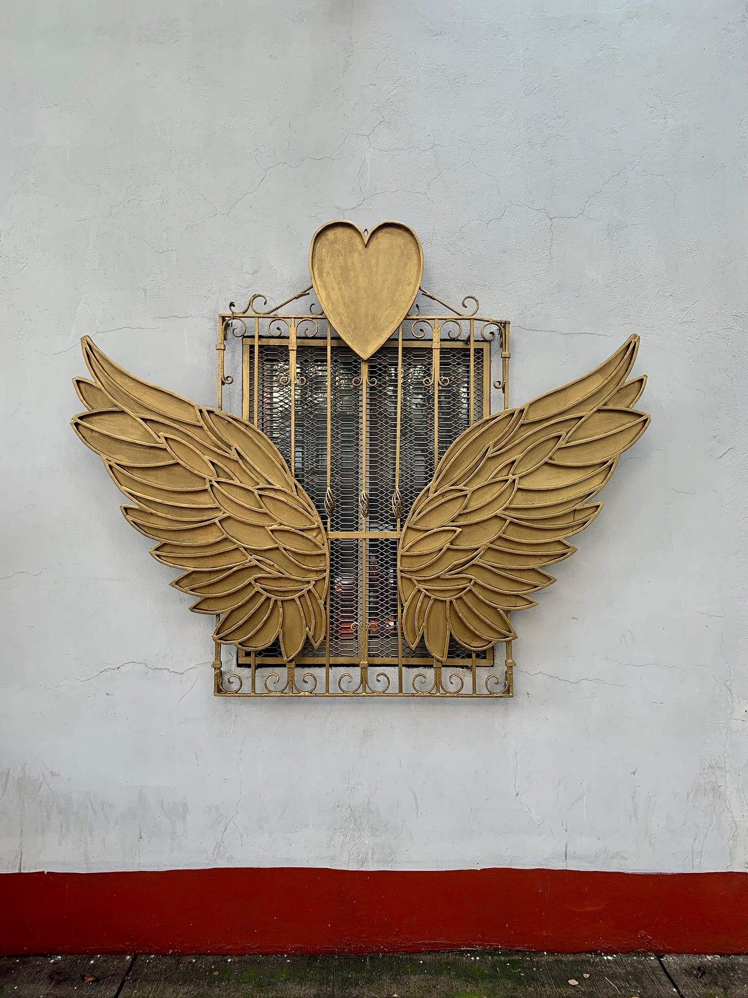 A window in Mexico City, the wall is white and the window has bars over it that are painted gold, with a heart at the top and gold wings spanning out from the middle. At the bottom is a red trim.