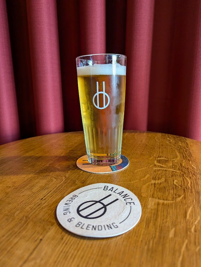 A 2/3 glass in Belgian gueuze style is full of golden saison beer. It has the Balance Brewing logo printed on it in white, and it sat on top of a wooden barrel that has been turned into a table, along with Balance Brewing beermats. Behind, a thick maroon curtain makes the beer feel like it's on a stage.