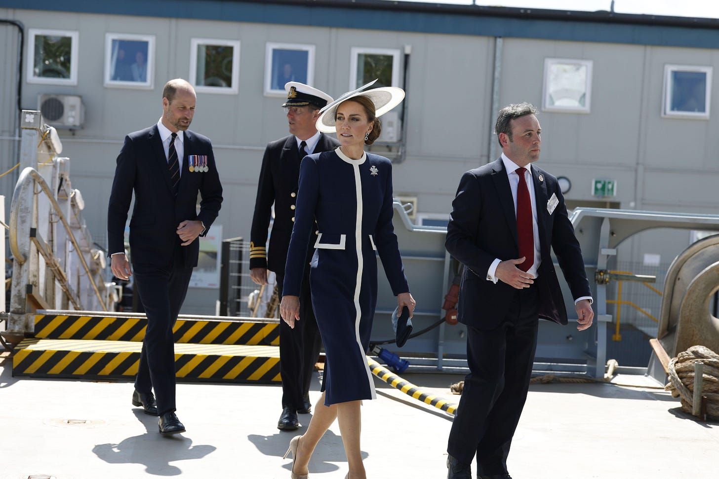 Princess Catherine walking in a navy blue dress and white hat, with Prince William following behind