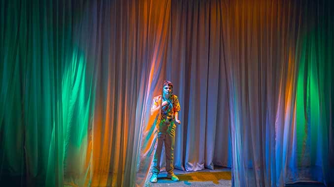 An actor stands defiantly center stage, flanked by curtains lit in greens and blues and oranges.