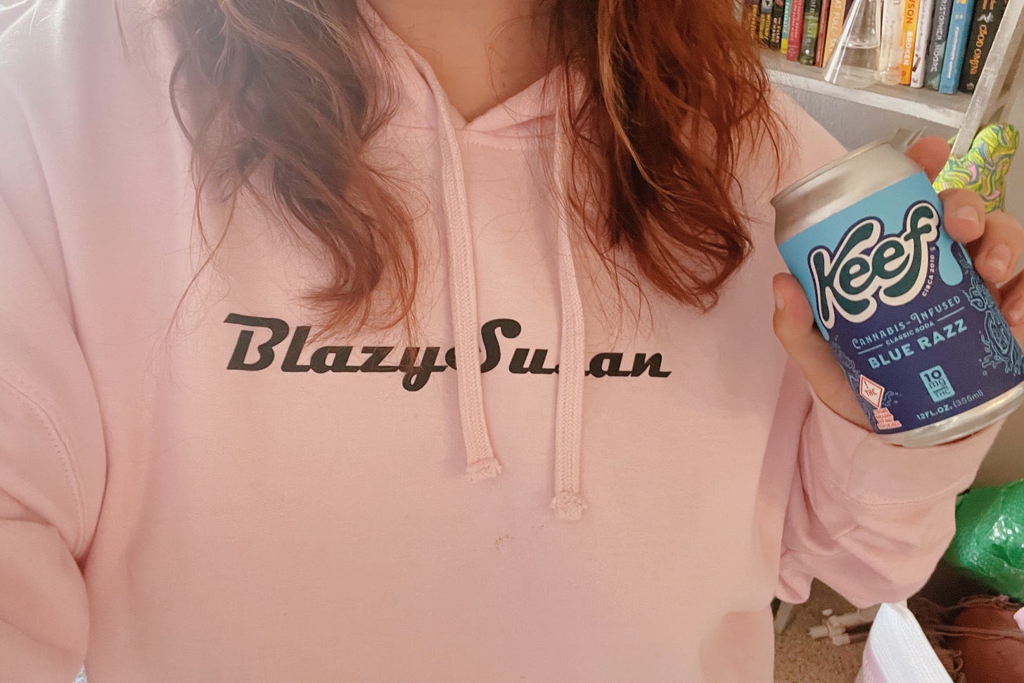 a torso with the head and neck just out of frame, wearing a pink hoodie that reads "Blazy Susan," holding the same can as in the previous picture