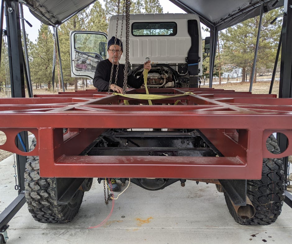 man stands outside next to a red subframe base on the back of the white work truck. It is a platform to hold the habitation box.