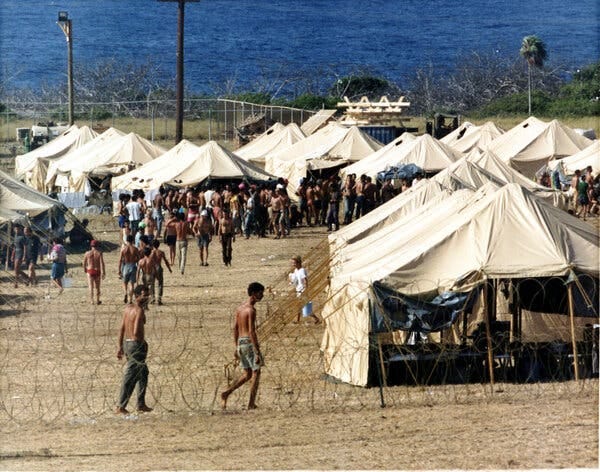 Shirtless men walking around a makeshift tent camp behind barbed wire in front of a bay.