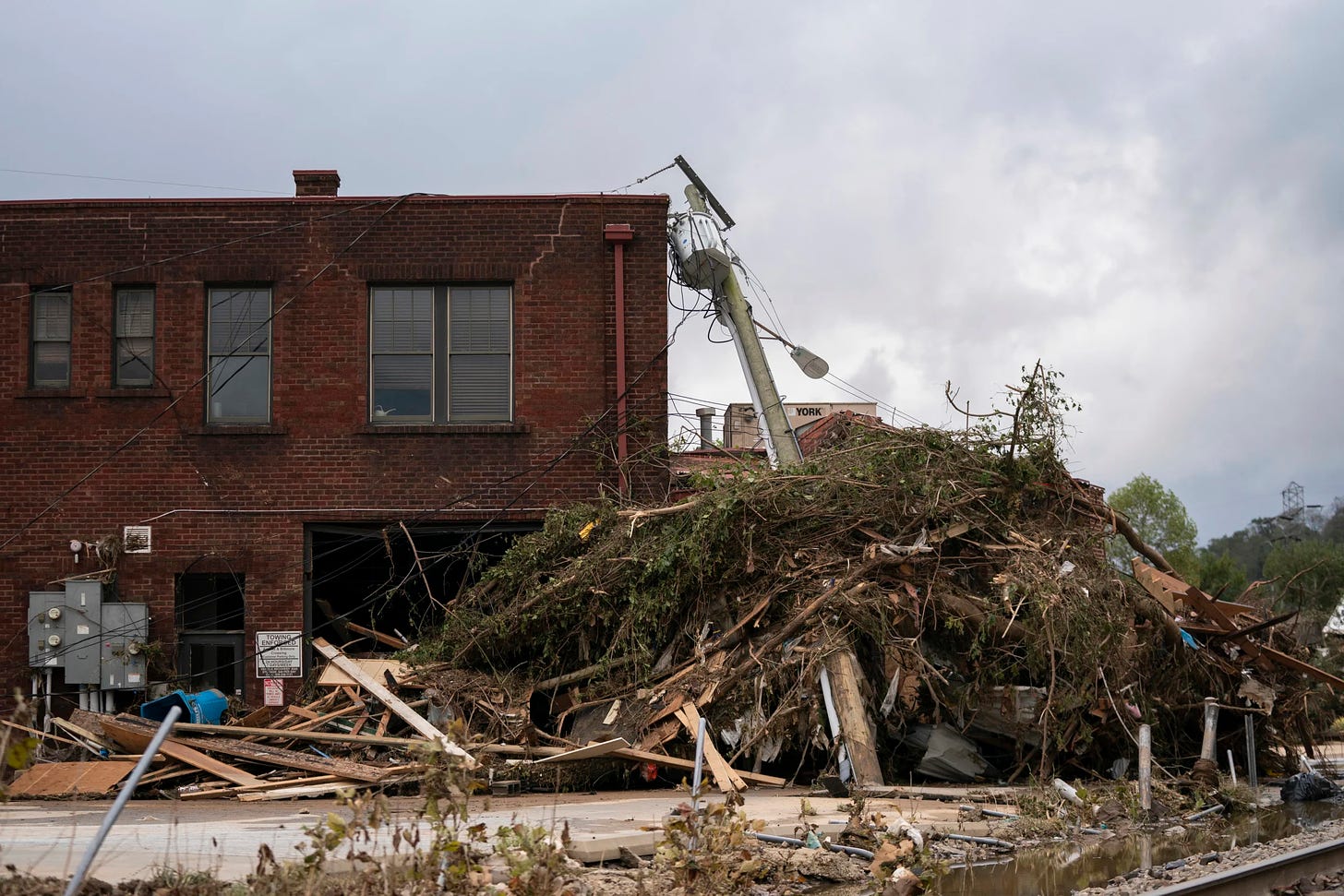 Debris from a destroyed building piled against a brick building after a natural disaster. Bent utility pole leaning on the debris. Overcast sky