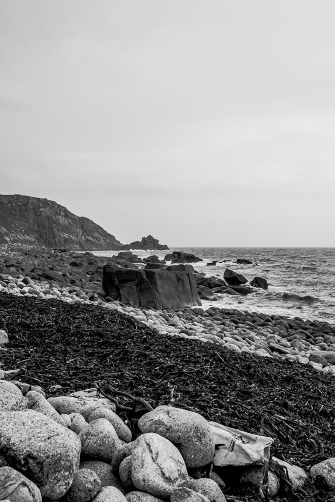 A black-and-white photo of a rocky beach with a thick line of seaweed. There are cliffs beyond and choppy waves in the sea.
