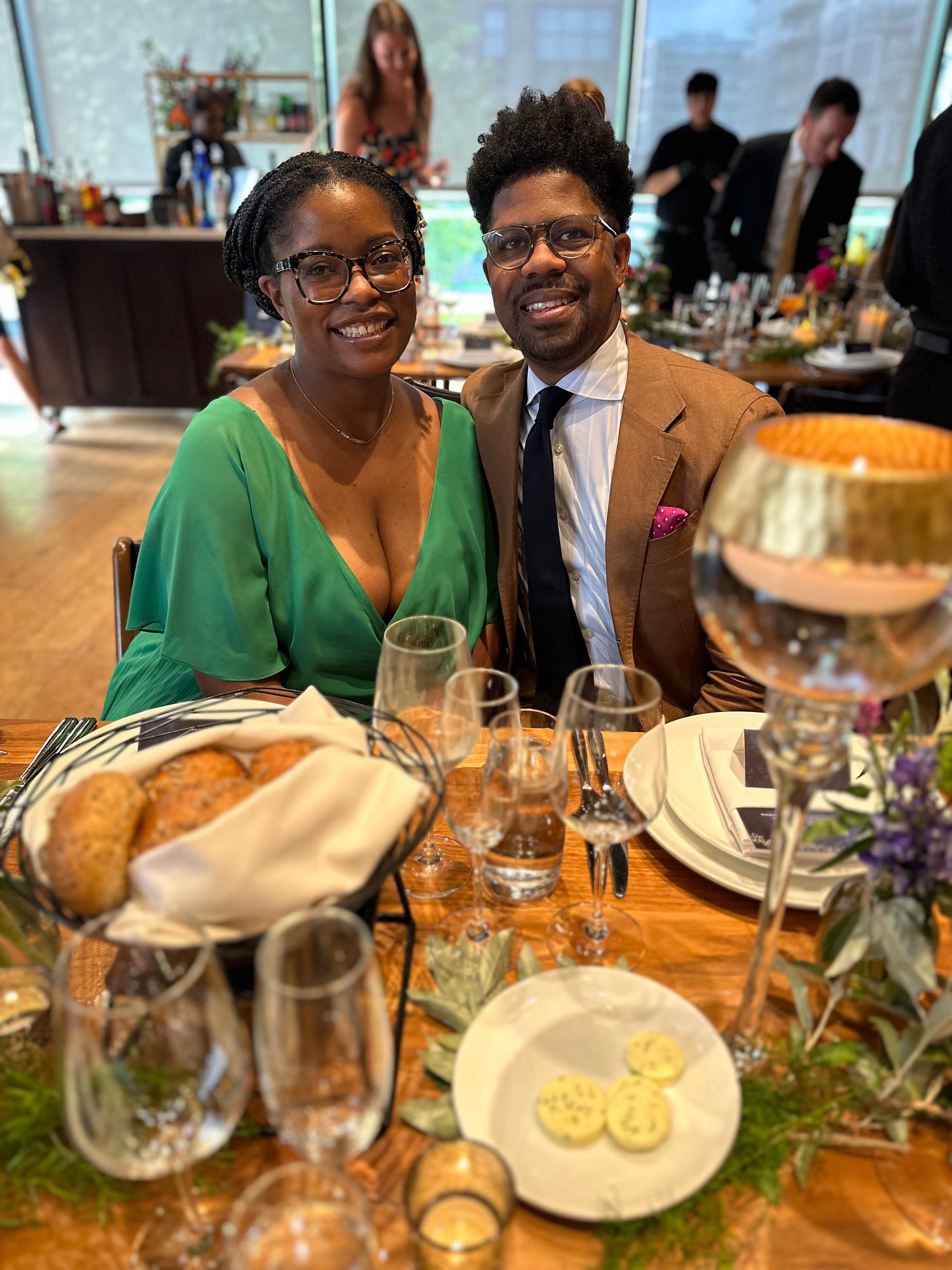 Picture of Black woman wearing a green dress and Black man wearing a brown suit at a wedding reception.