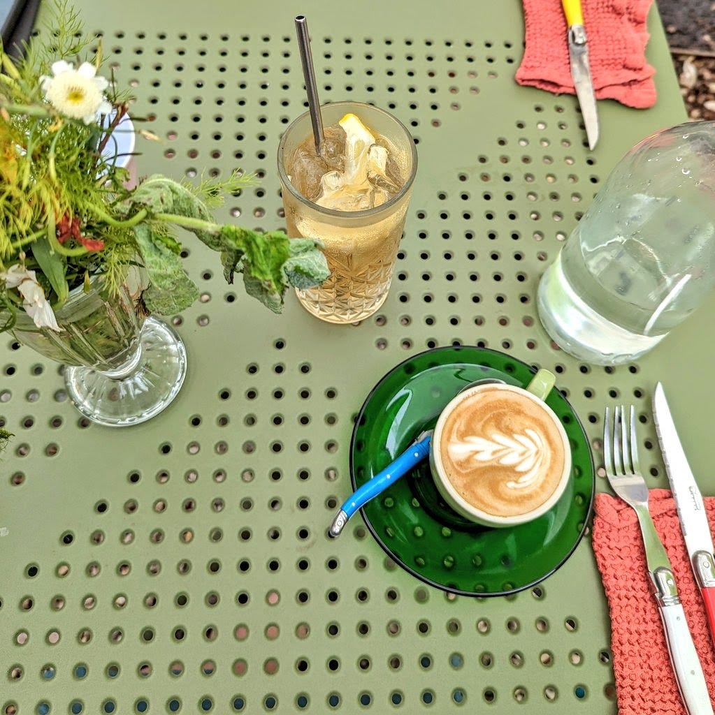 A flat white coffee and an iced tea on a green outdoor cafe table also featuring a vase of wildflowers, water jug, knives and forks 