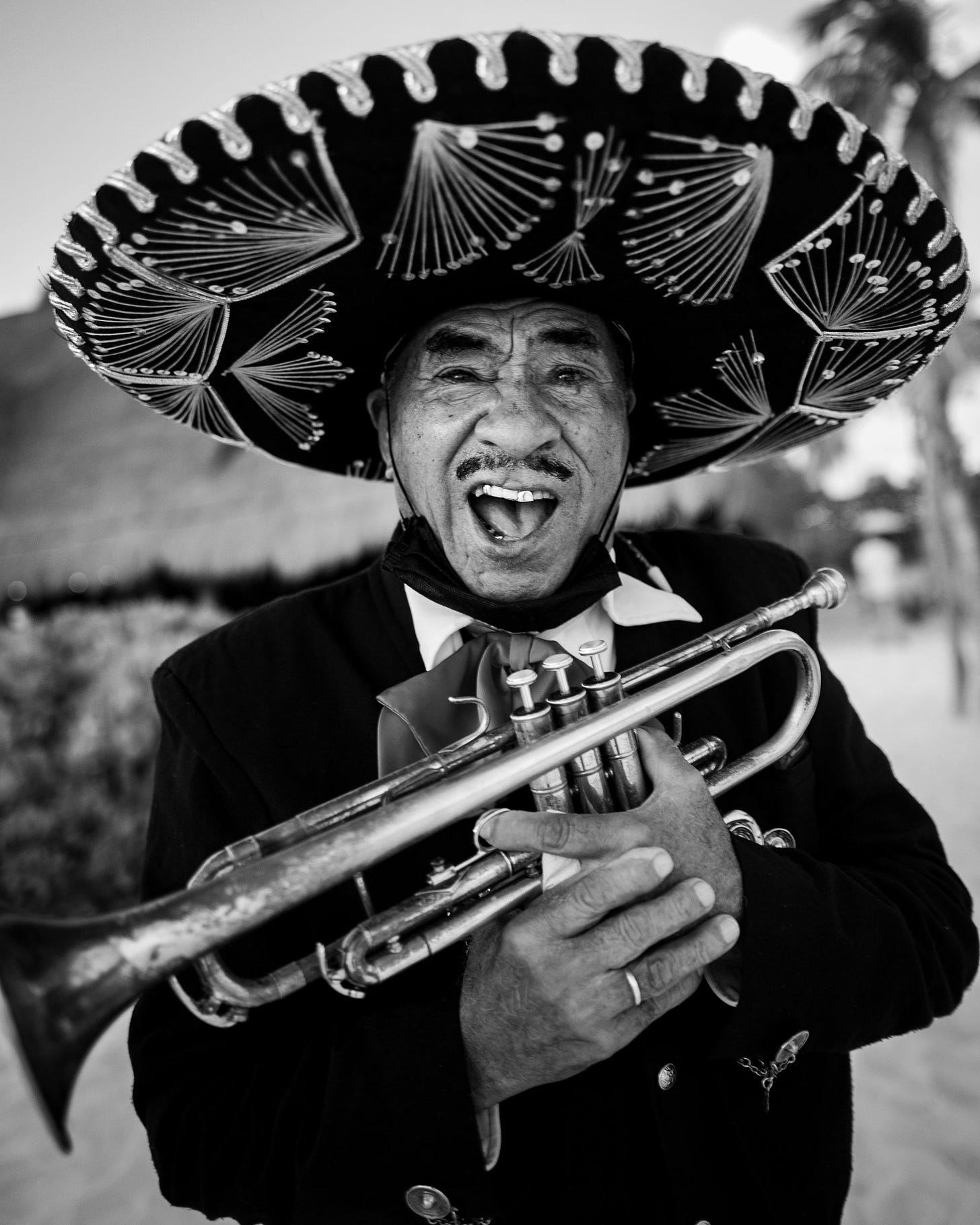 Close of a mariachi musician in black and white, holding a trumpet with an open mouth and an emotional or surprised look on his face.