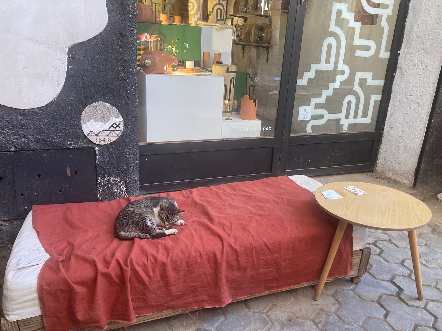A tabby cat with white points sleeps on a red draped bench outside of a shop