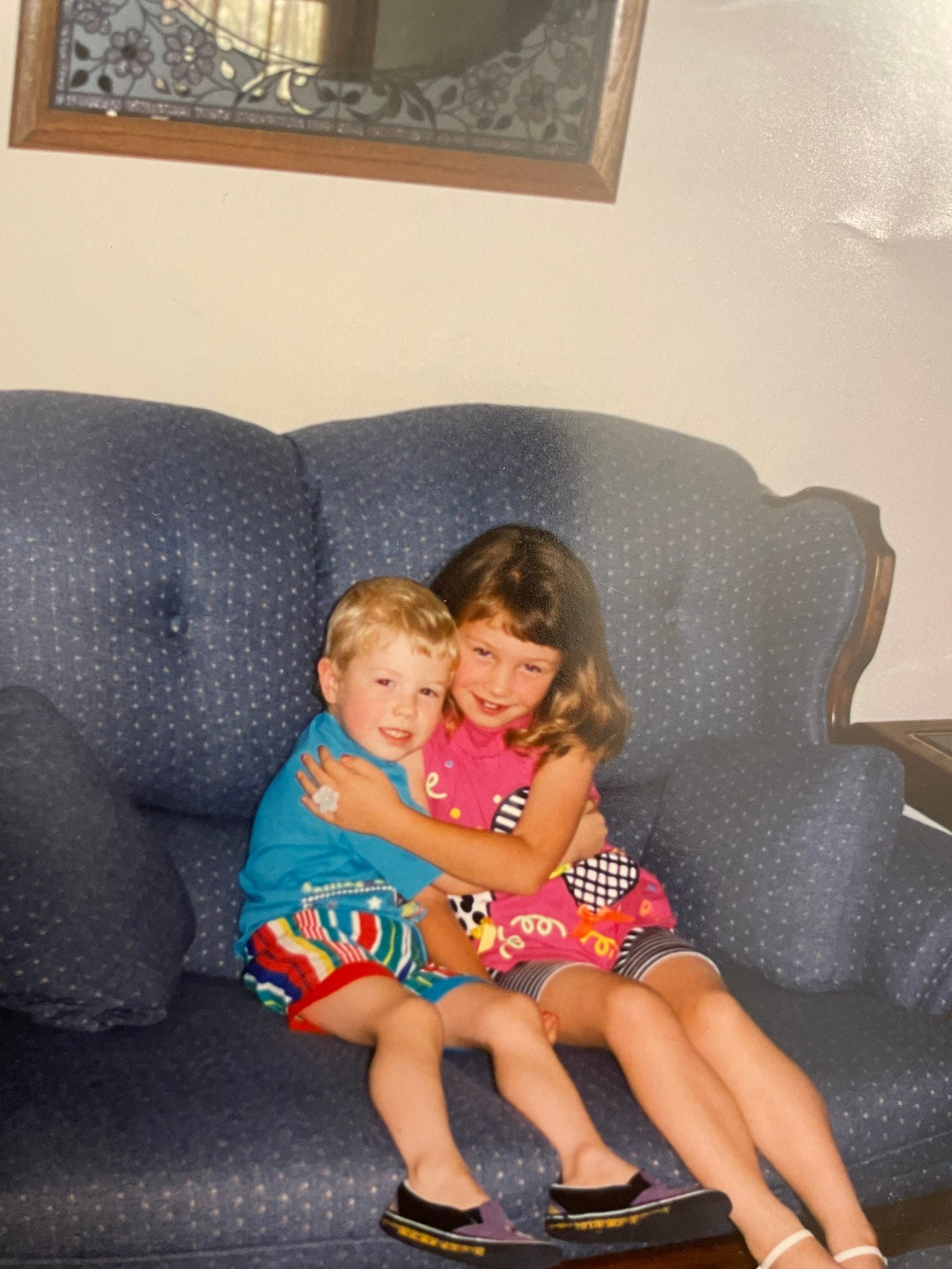 A young boy and girl are hugging each other, sitting on a blue couch, and smiling.