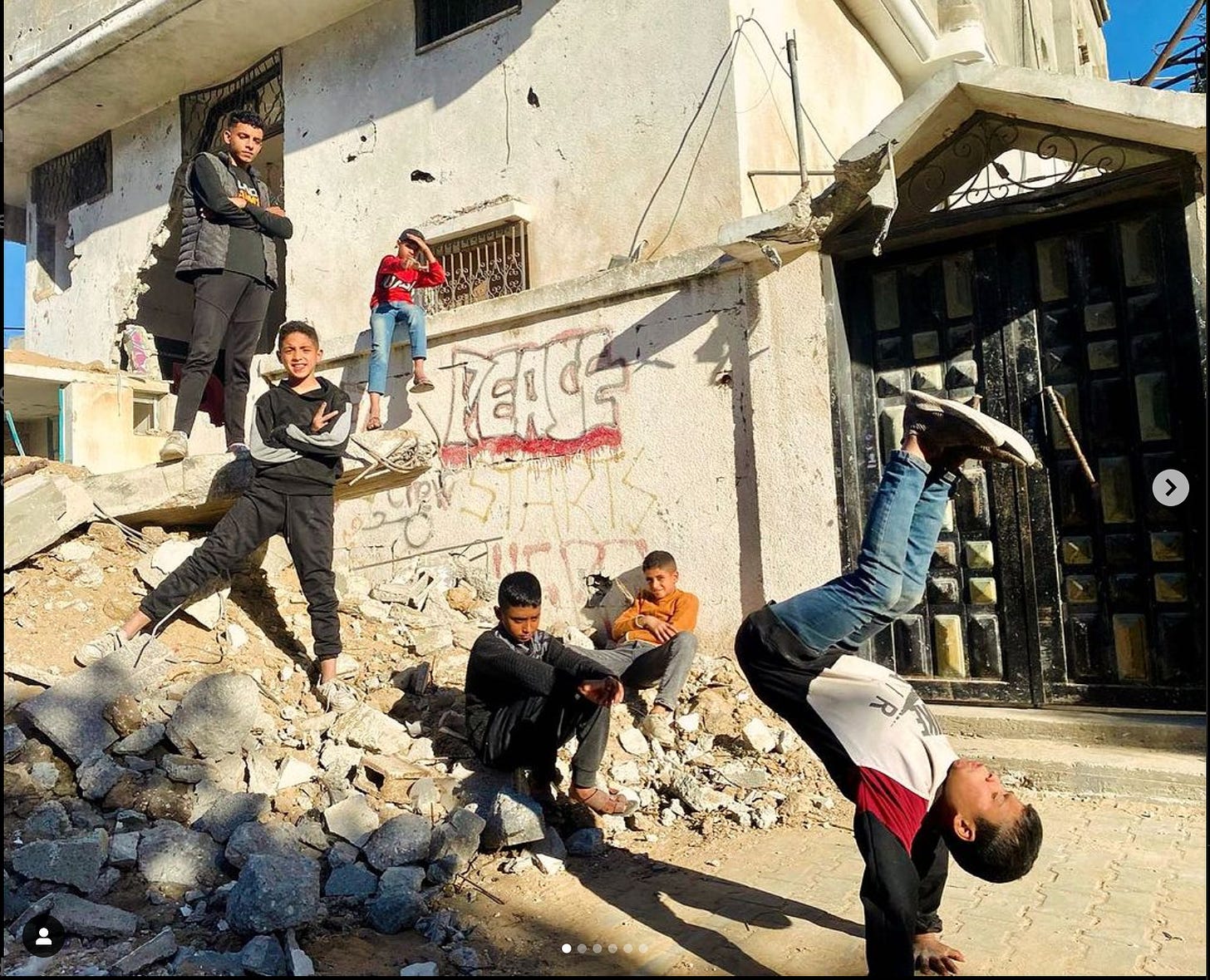 Young children pose and breakdance on and around rubble that's piled alongside a building with "peace" graffiti spray-painted on its walls.