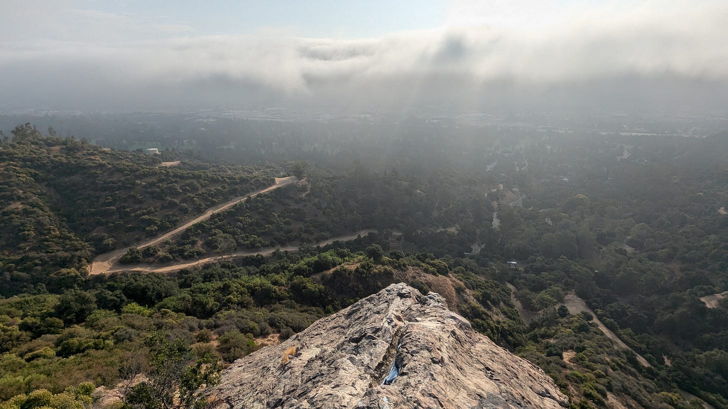 A dirt trail winds across hillsides covered in scrub, with a rock formation in the foreground, in dramatic early morning light