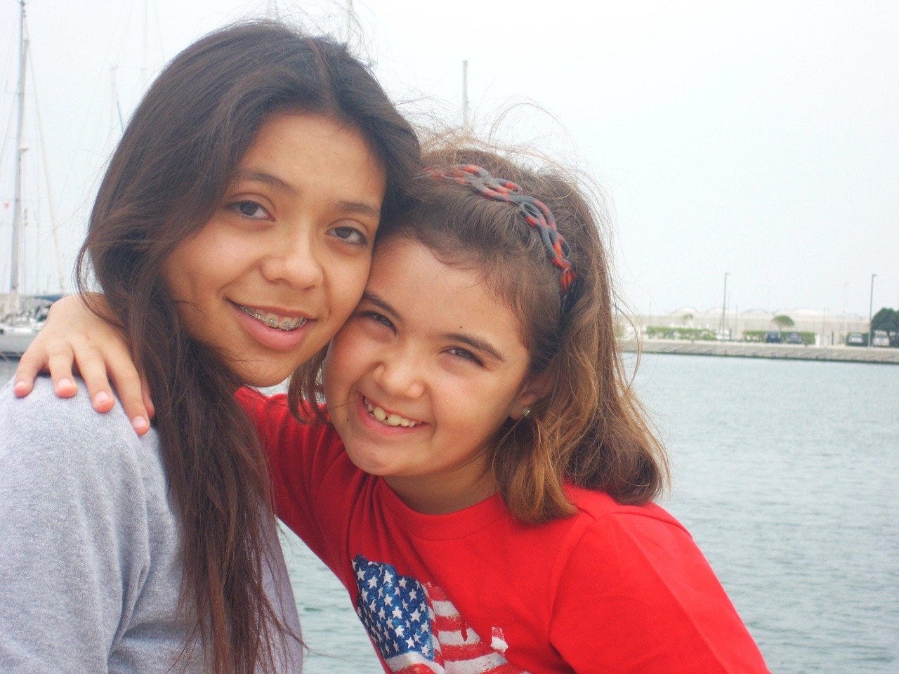 Two sisters embrace on a dock.  One has braces, the other is younger.