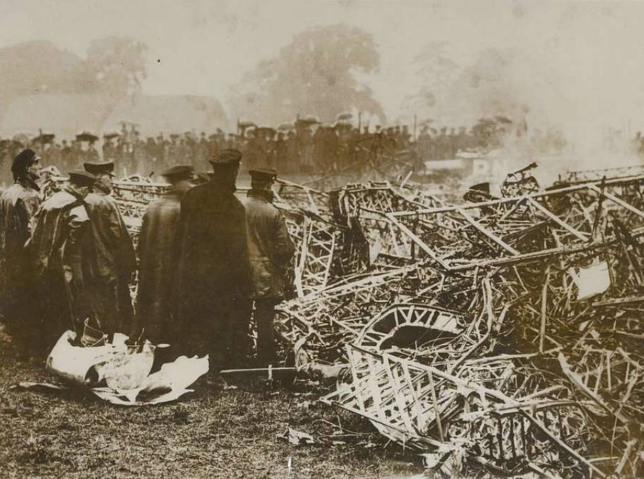 A black and white photo of a crowd of people, some of whom have umbrellas up, surrounding the mangled mess of Zeppelin L.31. The Zeppelin’s internal frame looks like an insurance “write-off” to me; far beyond the “it’ll buff out” stage.